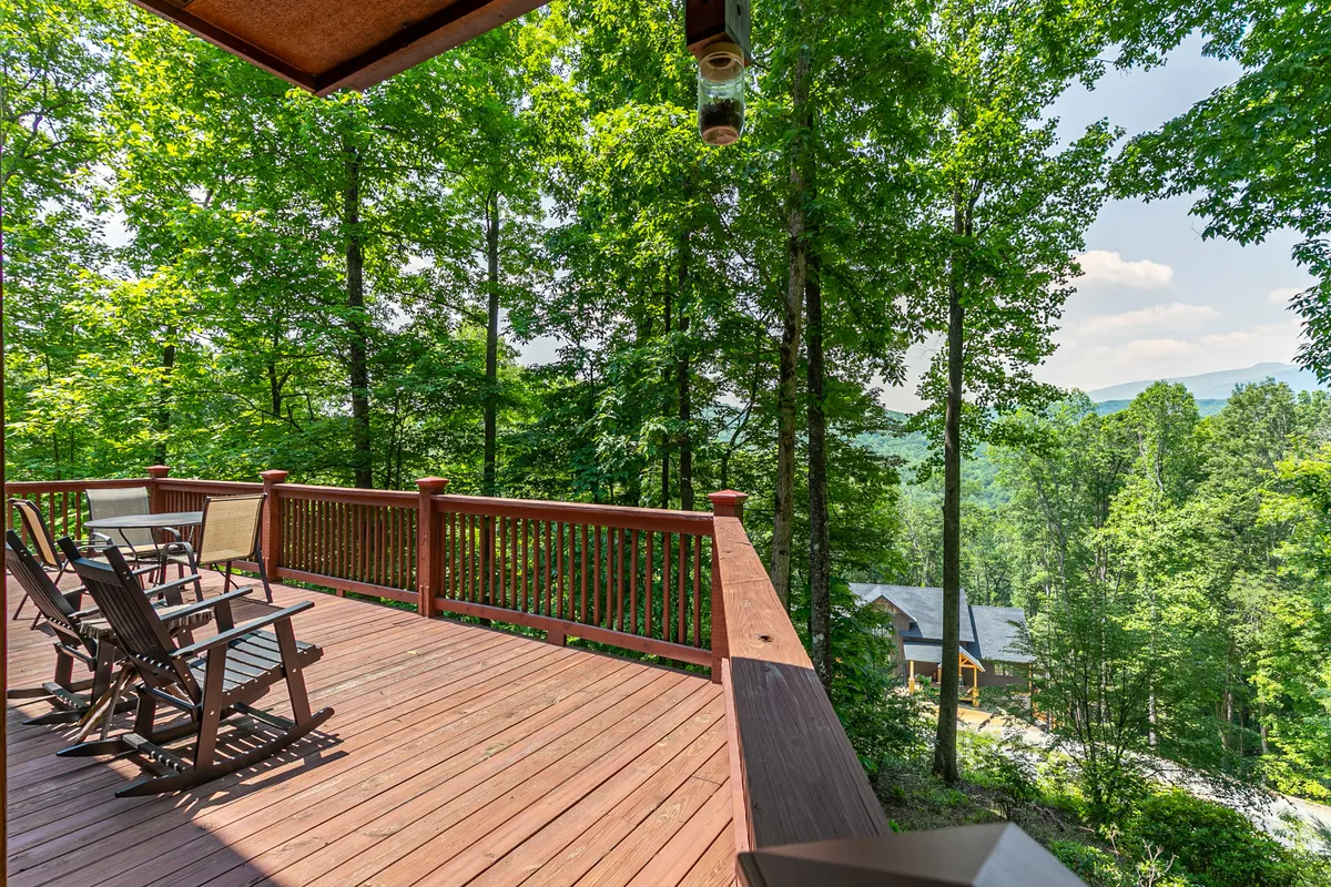 A spacious wooden deck with sturdy railings overlooks a stunning mountain view, surrounded by tall trees. The deck is furnished with seating, making it a comfortable spot for relaxing or enjoying a meal outdoors while soaking in the natural beauty.