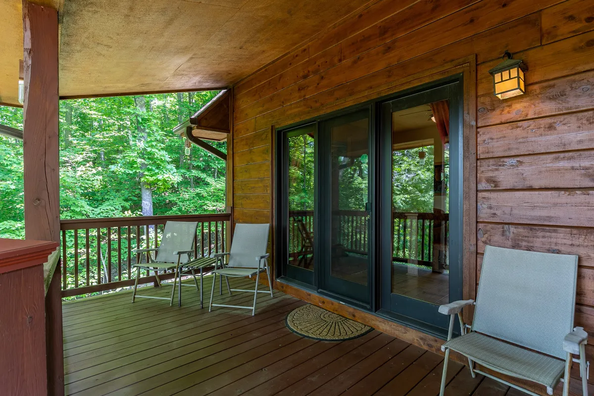A peaceful wooden deck bordered by green foliage, with outdoor chairs and sliding glass doors connecting the deck to the cabin’s interior. The scene is serene, inviting guests to relax outside, socialize, or enjoy the mountain air under the open sky.