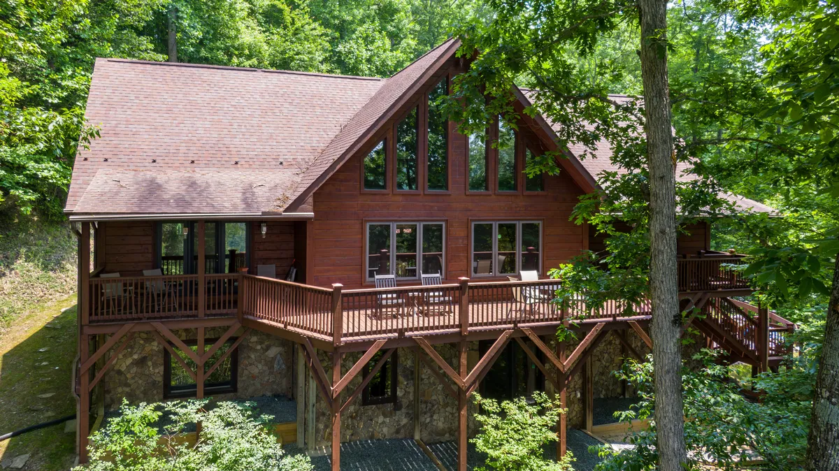The hero image showcases the inviting log cabin nestled among lush trees, with expansive windows reflecting the golden sunset. A spacious wooden deck wraps around the cabin, set atop a sturdy stone foundation, offering ample space for outdoor dining, relaxing with coffee at sunrise, or stargazing in the tranquil mountain air.