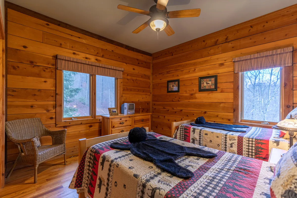A cozy bedroom with twin beds adorned with patterned quilts, natural wooden walls, a wicker chair, and large windows letting in abundant light and offering relaxing views of the outdoors.