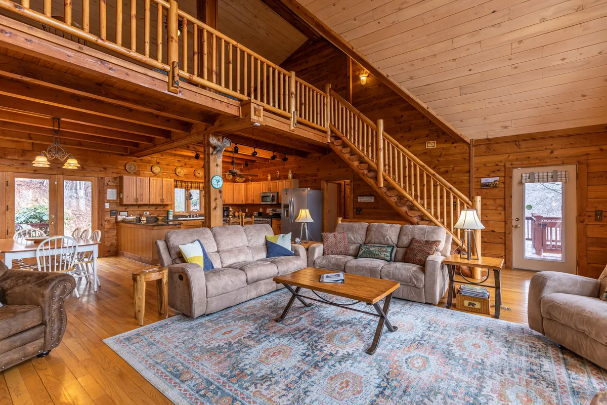 The photo showcases the heart of the home, with a cozy seating area arranged around a central coffee table, a staircase leading upwards, and a view into the kitchen and dining zones. The fireplace and intimate arrangement signal a space ideal for gatherings, while the kitchen and dining table in the background reinforce the open-concept design.