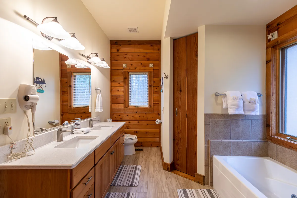 A calm and spacious bathroom with a double vanity, large bathtub perfect for soaking, wooden accents, big mirrors, and plenty of towels. The room is softly lit and designed for relaxation and self-care.