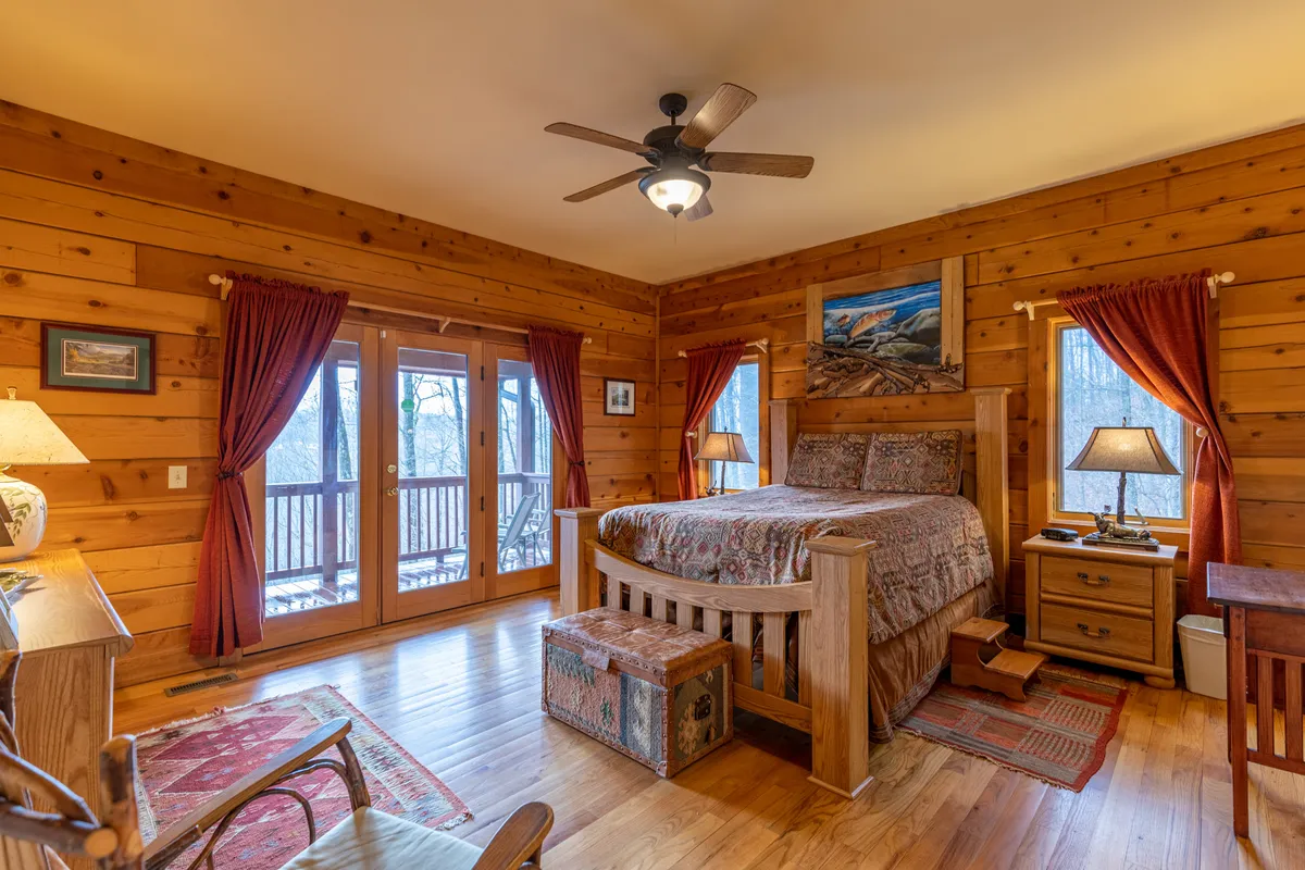 A tranquil bedroom featuring a comfortable bed, French doors that open to natural views, tasteful artwork adorning the wall, soft lamps, and a plush rug. The room exudes warmth and invites relaxation, with ample daylight streaming through the glass doors.