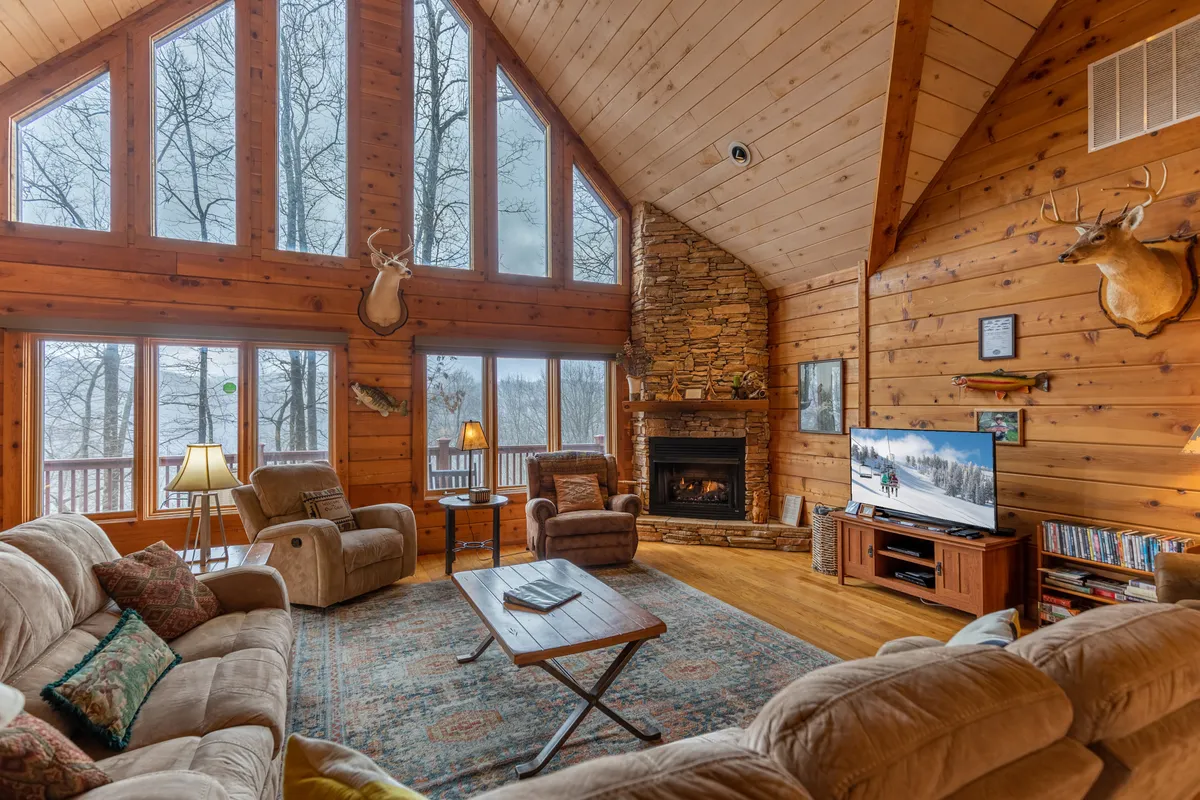 A spacious living area with large windows letting in natural light and offering a glimpse of the outdoors. The room features a striking stone fireplace, comfortable seating, and decorative touches like mounted deer heads, blending rustic charm with modern comfort. This is a communal space ideal for gathering after a day of exploring the town.