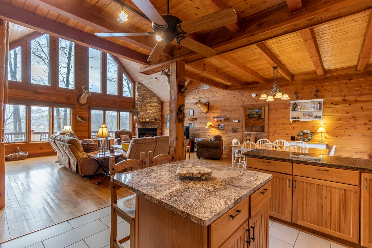 This image captures a spacious open-plan living area, featuring large windows that invite abundant natural light and frame sweeping mountain views. A prominent stone fireplace creates a cozy focal point, while a kitchen island nearby hints at seamless flow between the living, cooking, and dining spaces. Wooden ceiling beams and warm tones enhance the sense of comfort and togetherness.