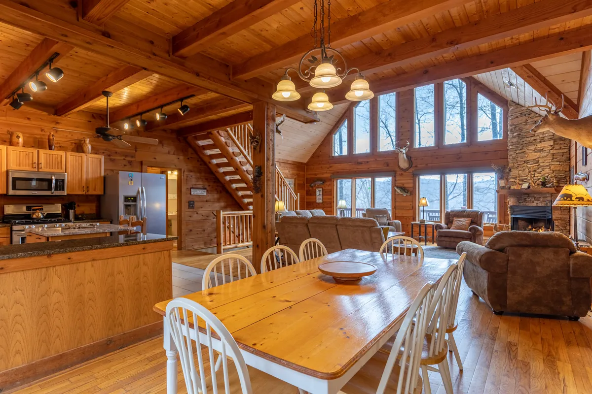 A warm, family-friendly dining area with a sturdy wooden table sits adjacent to a stone fireplace, surrounded by large windows offering picturesque views of nature. Comfortable seating and rustic decor create an inviting ambiance for shared meals and special occasions.