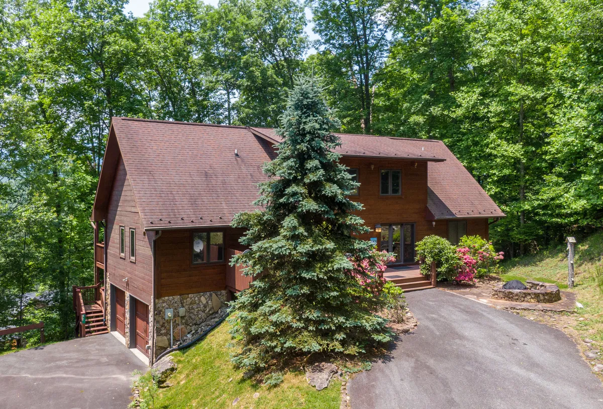 The wooden exterior of the lodge is surrounded by lush greenery, flowering shrubs, and a stone pathway leading up to a welcoming porch, blending the home beautifully into its natural environment. The image evokes tranquility and the spirit of outdoor living.